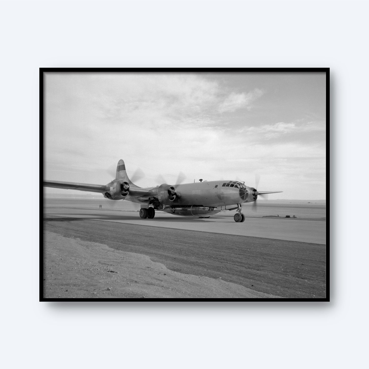 A black and white photograph from the "B-29 Mothership Aircraft Print" collection showcases a vintage Boeing B-29 Superfortress on a runway. The iconic plane, equipped with four propeller engines, is captured from the front-left side against a backdrop of a cloudy sky and an expansive, barren landscape.