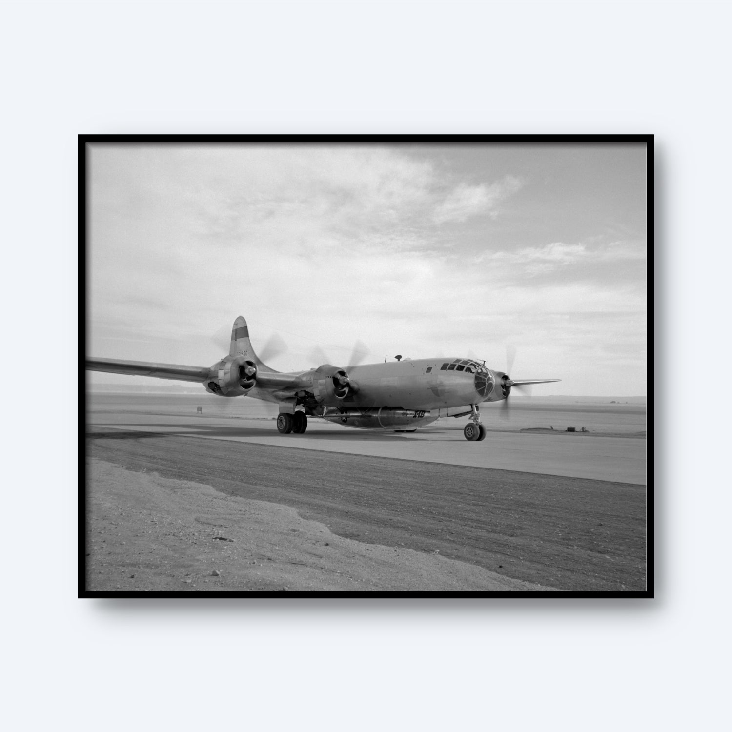 A black and white photograph from the "B-29 Mothership Aircraft Print" collection showcases a vintage Boeing B-29 Superfortress on a runway. The iconic plane, equipped with four propeller engines, is captured from the front-left side against a backdrop of a cloudy sky and an expansive, barren landscape.