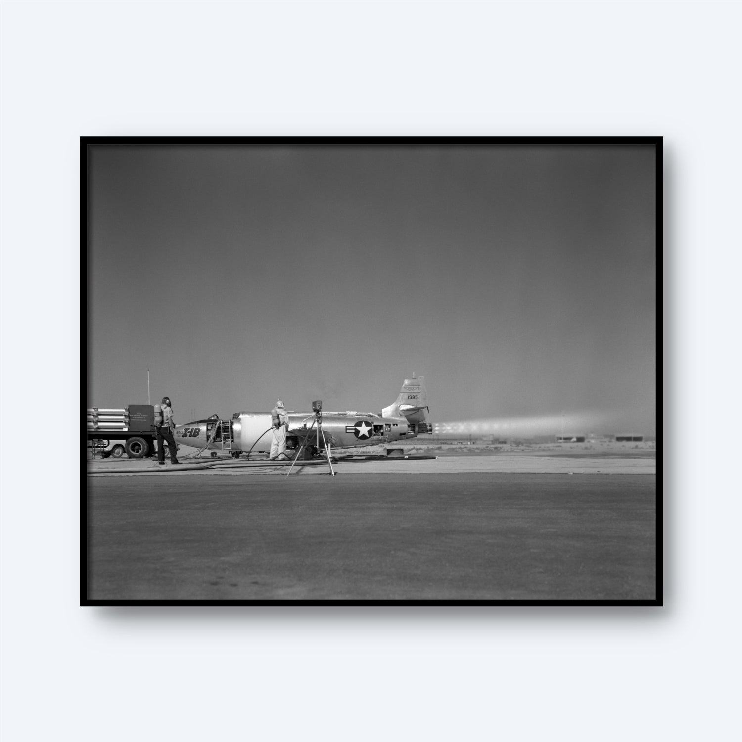A black-and-white image depicts the X-1B Jet Engine parked on a runway. Two military personnel are working around the aircraft, one near the tail and the other near the front, surrounded by equipment and a truck. Capturing this piece of aerospace history, this stunning wall art portrays a bright, clear day.