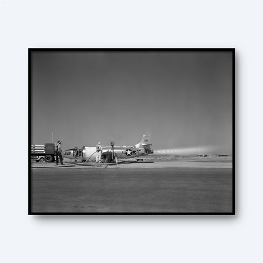 A black-and-white image depicts the X-1B Jet Engine parked on a runway. Two military personnel are working around the aircraft, one near the tail and the other near the front, surrounded by equipment and a truck. Capturing this piece of aerospace history, this stunning wall art portrays a bright, clear day.