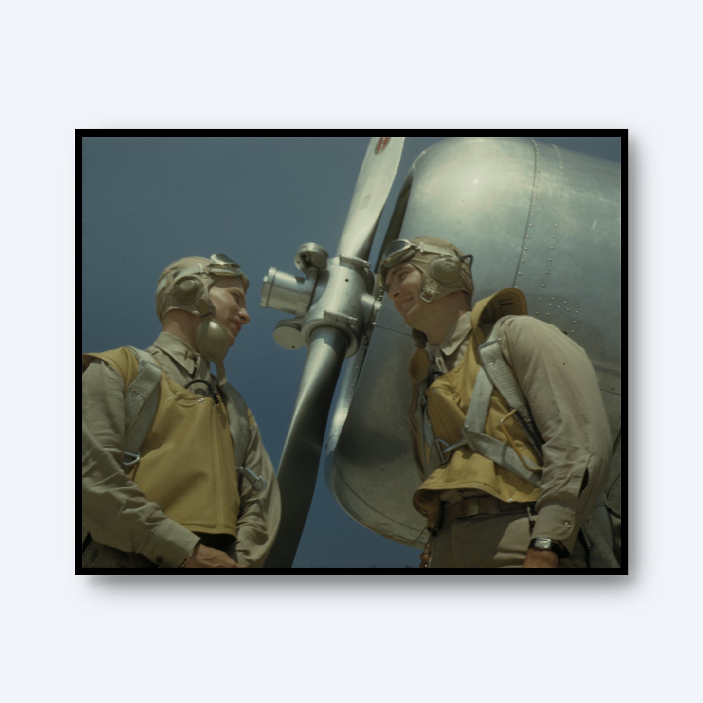 The "WW2 Pilots Print" captures two World War II pilots clad in tan flight suits and helmets with goggles, standing beside an airplane propeller. They are engaged in a joyful conversation against a clear blue sky, with the metallic body of their aircraft gleaming in the background, making it a timeless piece of historical wall art.