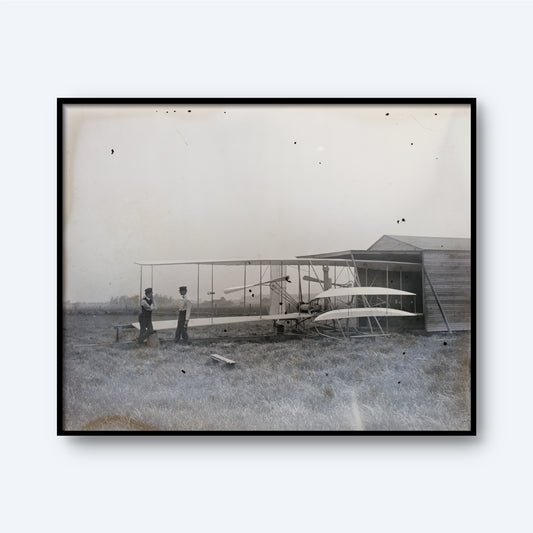 A black-and-white photograph titled "The Wright Brothers Print," depicting an early airplane with a biplane structure and twin wings, standing outside a small hangar. Two individuals are positioned near the aircraft, one on each side. This piece of Early Aviation Art is set in a grassy field under an overcast sky.