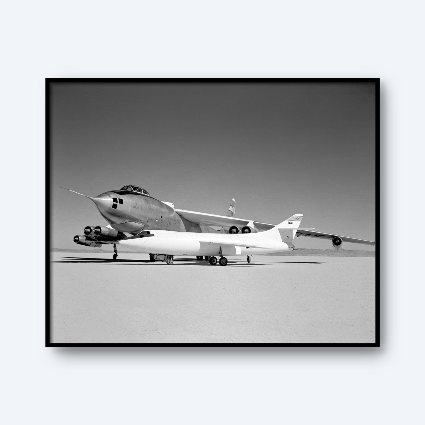 Monochromatic image capturing a significant moment in aviation history, featuring a large Boeing B-47A Stratojet with a smaller Douglas D-558-II attached beneath its fuselage. The aircraft are stationary on a dry, flat surface reminiscent of a desert or test area, emphasizing their sleek and sharp-nosed design. This print is titled "Douglas D-558-II and Boeing B-47A Stratojet Print | X Planes.