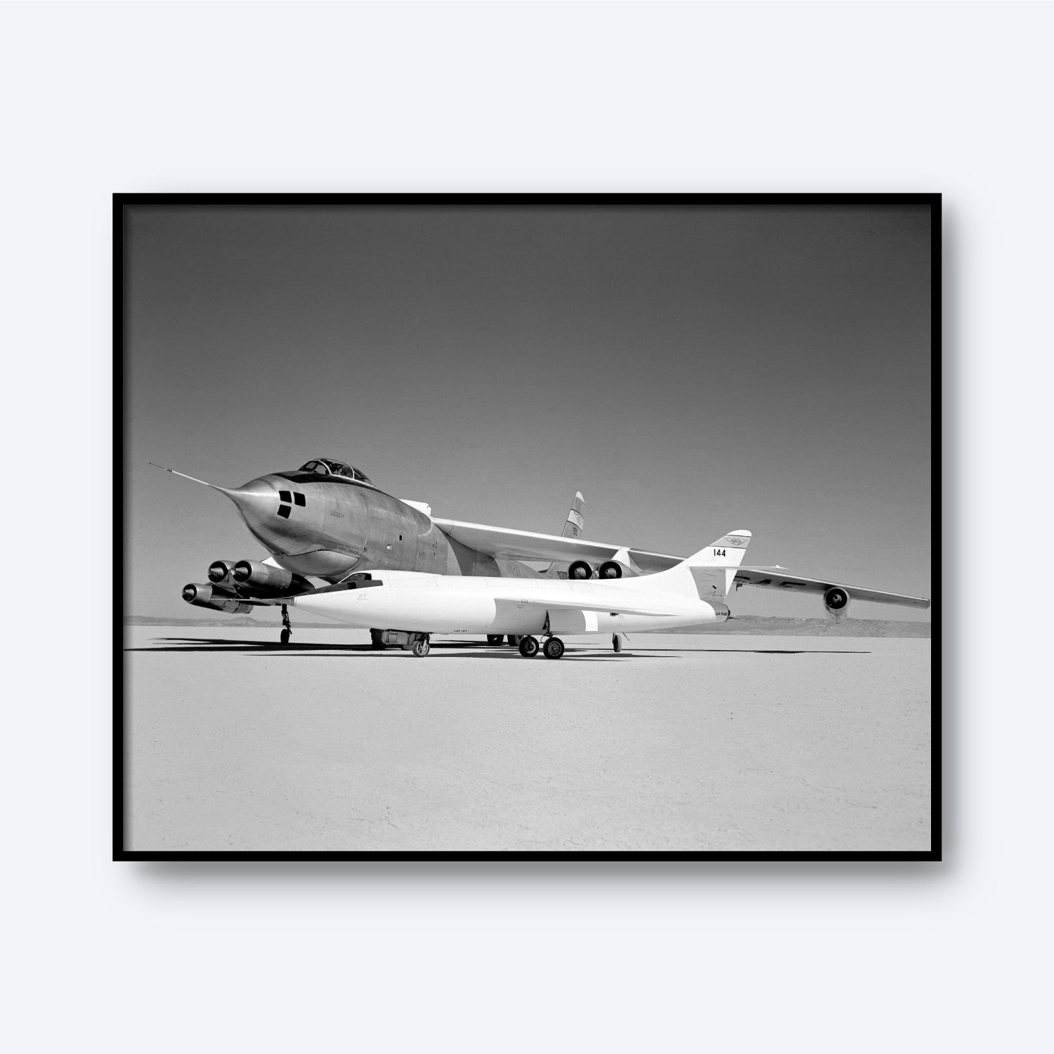Monochromatic image capturing a significant moment in aviation history, featuring a large Boeing B-47A Stratojet with a smaller Douglas D-558-II attached beneath its fuselage. The aircraft are stationary on a dry, flat surface reminiscent of a desert or test area, emphasizing their sleek and sharp-nosed design. This print is titled "Douglas D-558-II and Boeing B-47A Stratojet Print | X Planes.