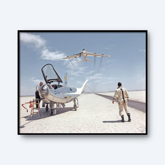 The HL-10 on Lakebed with B-52 flyby Print showcases a futuristic lifting body aircraft featuring an open cockpit, resting on the dry, cracked surface of a desert lakebed. Two individuals are nearby; one stands on a step ladder while the other approaches from the ground. Above them, a small B-52 soars across the clear blue sky.