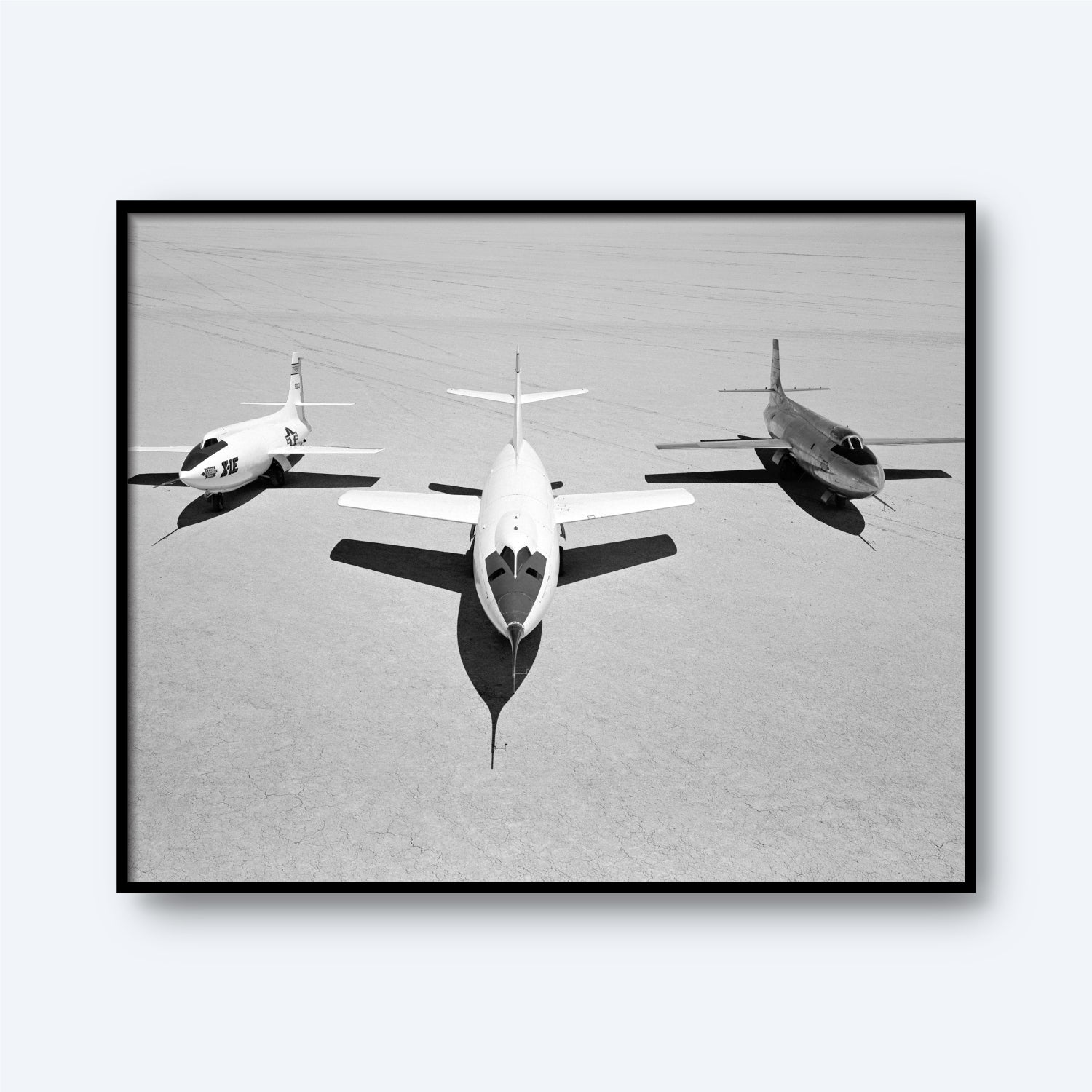 A black-and-white photo captures three military jets aligned side by side on a vast, barren lakebed. The central jet, reminiscent of the sleek X-1B design, is positioned slightly ahead of its two flanking companions [D-558-II and X-1E]. The ground appears flat and desolate under a stark sky.