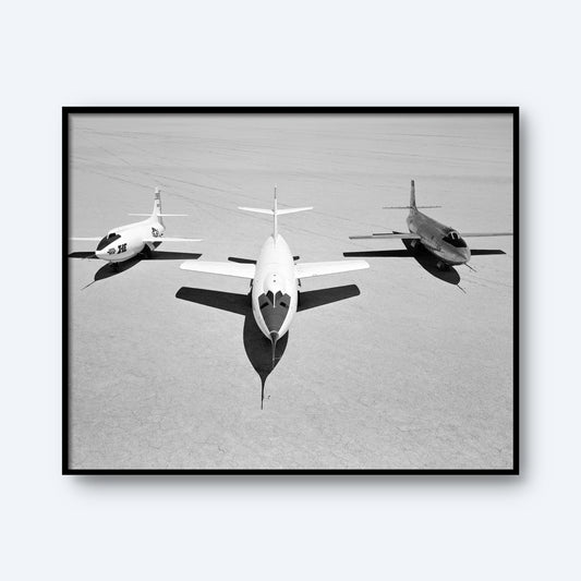 A black-and-white photo captures three military jets aligned side by side on a vast, barren lakebed. The central jet, reminiscent of the sleek X-1B design, is positioned slightly ahead of its two flanking companions [D-558-II and X-1E]. The ground appears flat and desolate under a stark sky.