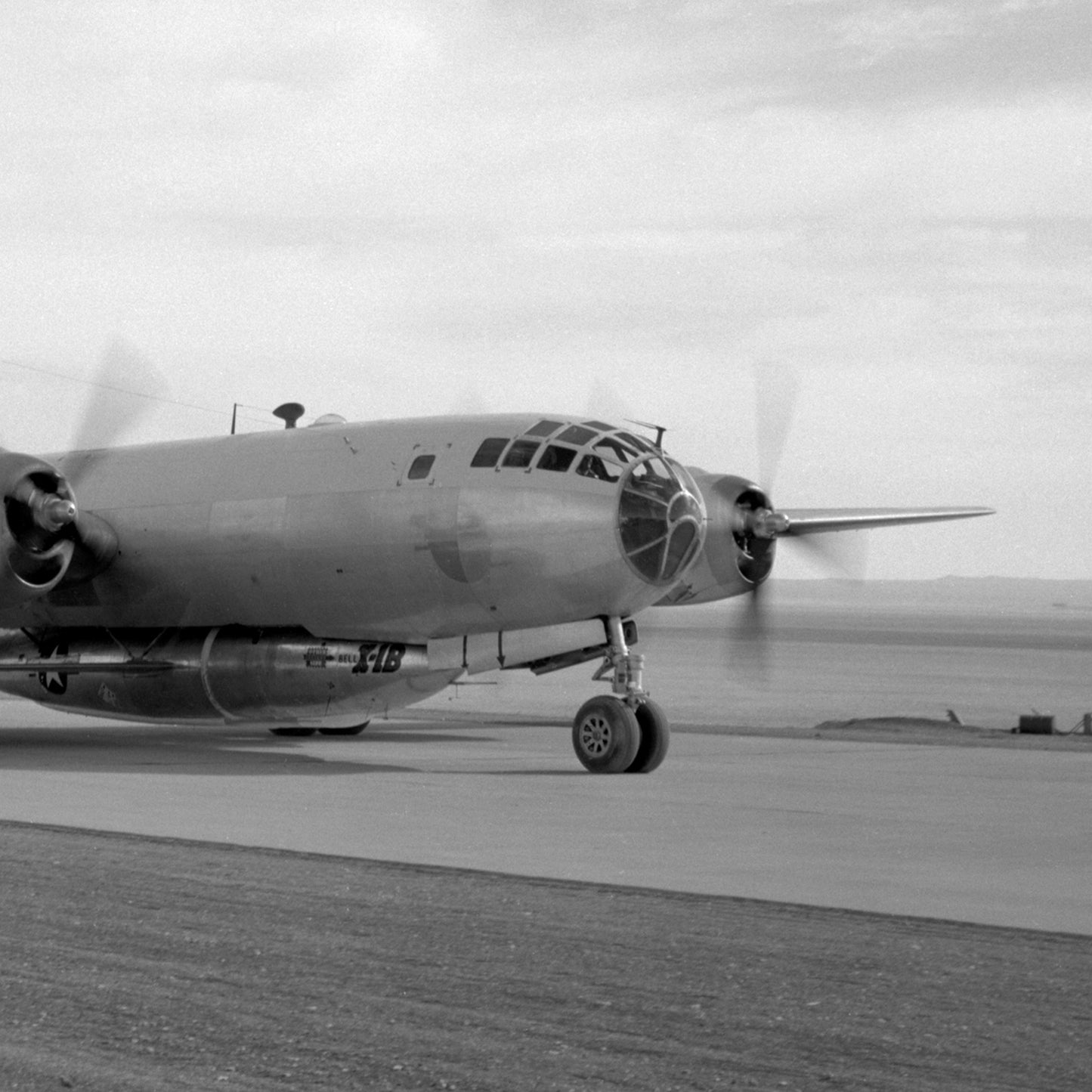 A black-and-white photo of the B-29 Mothership Aircraft Print shows a large propeller-driven military aircraft on the tarmac. The Boeing B-29 Superfortress, recognizable by its two propellers on each wing, showcases a glass cockpit and extended landing gear. The spinning propellers indicate the engines are running, set against an open landscape background.