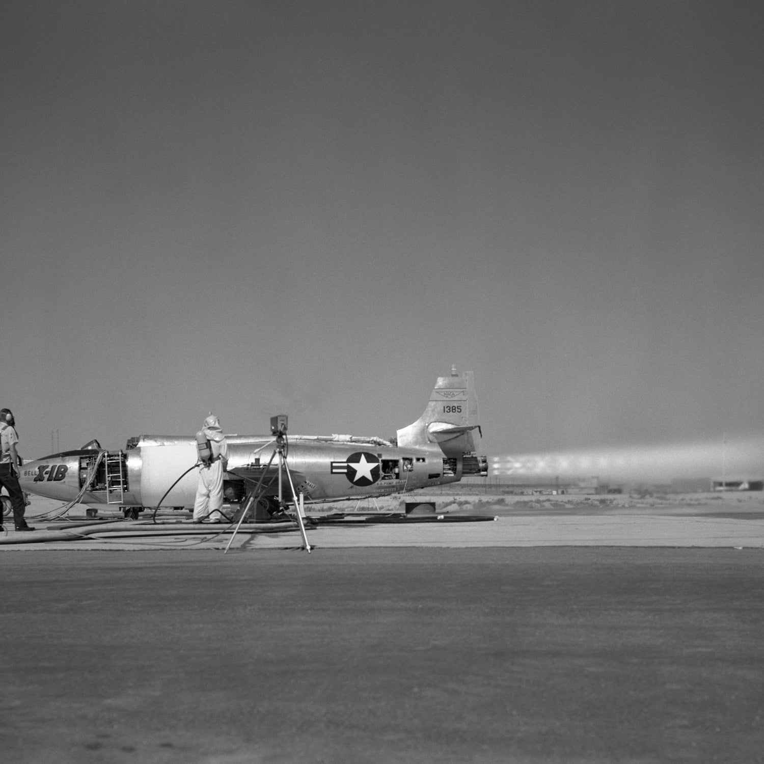 Black and white image of the vintage X-1B Jet Engine parked on an airfield. Two people are seen near it, one of whom appears to be working or inspecting the aircraft. The jet, featuring a USAF insignia, has a vapor trail behind it. A tripod is set up in front, capturing this piece of aerospace history for wall art.