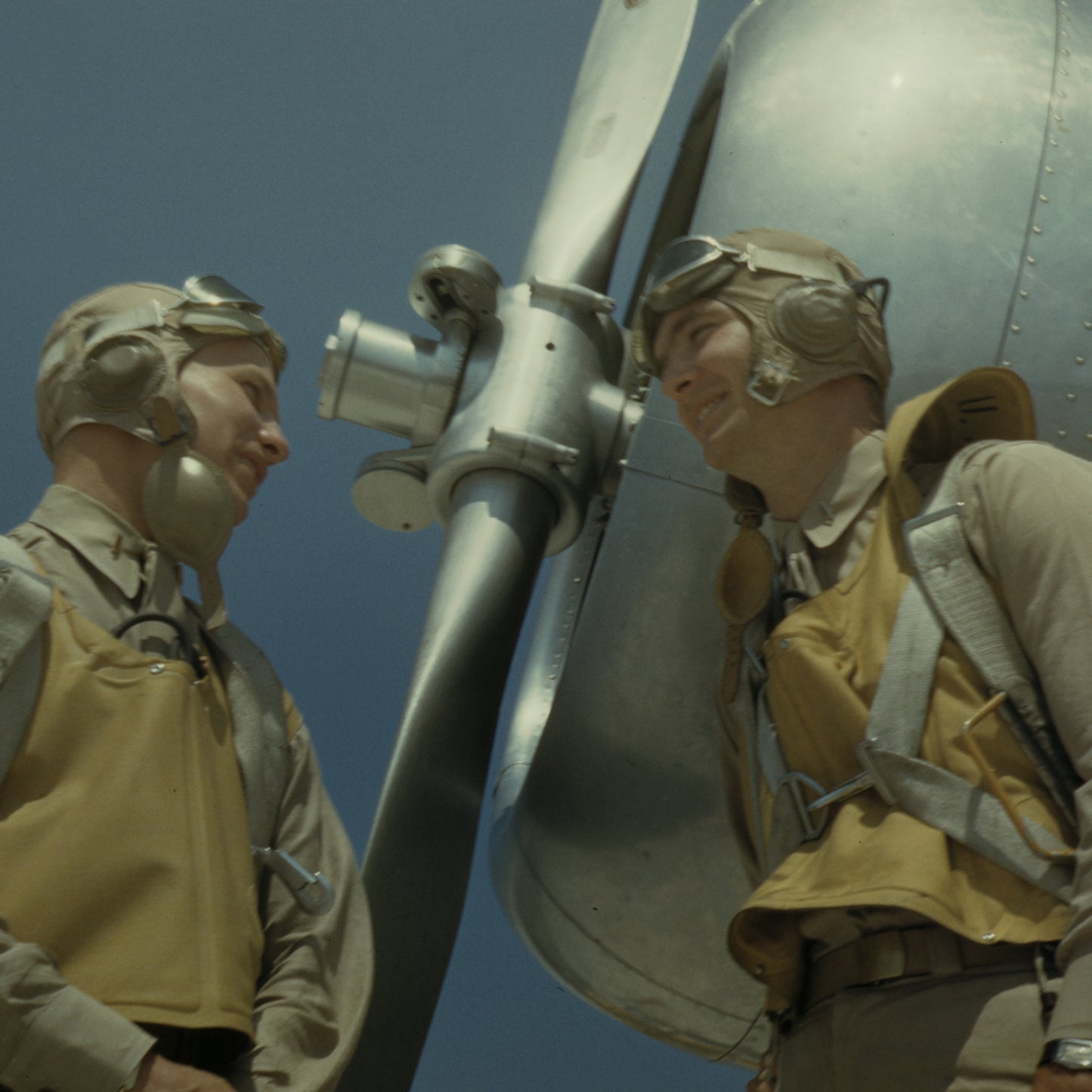 Two World War II pilots dressed in vintage flight gear, such as helmets, goggles, and life vests, stand beside an aircraft propeller, smiling and engaging in conversation. The clear blue sky hints at a sunny day—an ideal setting for this moment captured perfectly as the WW2 Pilots Print historical wall art.