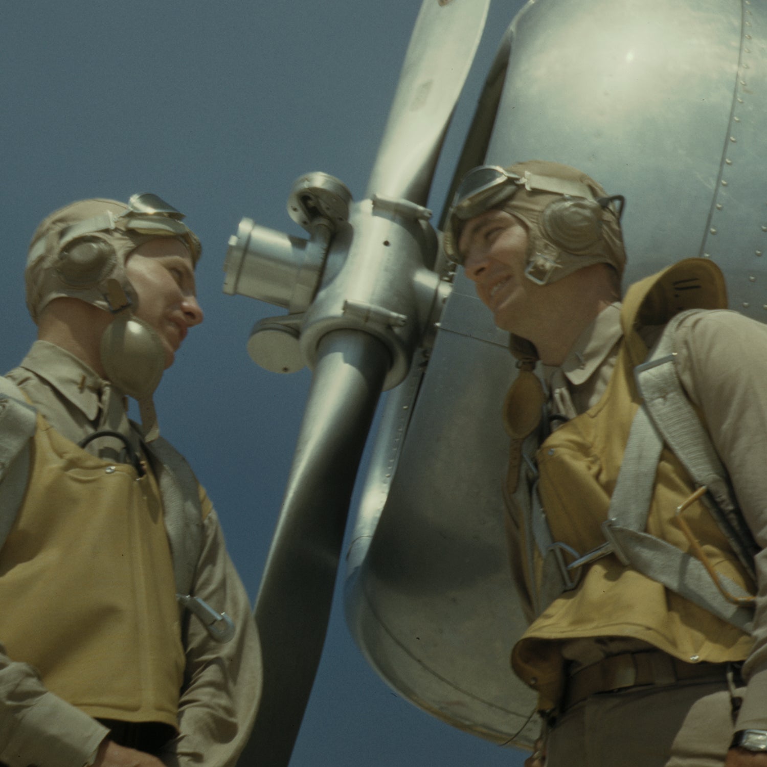 Two World War II pilots dressed in vintage flight gear, such as helmets, goggles, and life vests, stand beside an aircraft propeller, smiling and engaging in conversation. The clear blue sky hints at a sunny day—an ideal setting for this moment captured perfectly as the WW2 Pilots Print historical wall art.