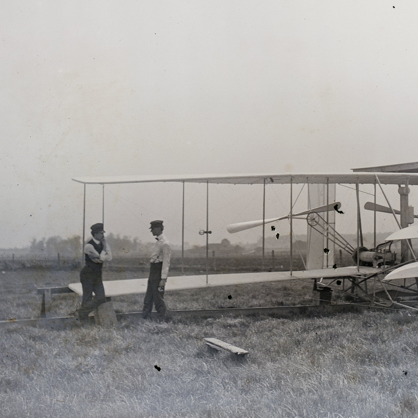 Two men stand in front of a vintage biplane on a grassy field, evoking Early Aviation Art. The man on the left leans against the aircraft, while the man on the right stands upright, both gazing at the plane. The background is a foggy open space with a distant tree line, making it perfect for Historical Wall Decor. This scene is depicted in "The Wright Brothers Print.