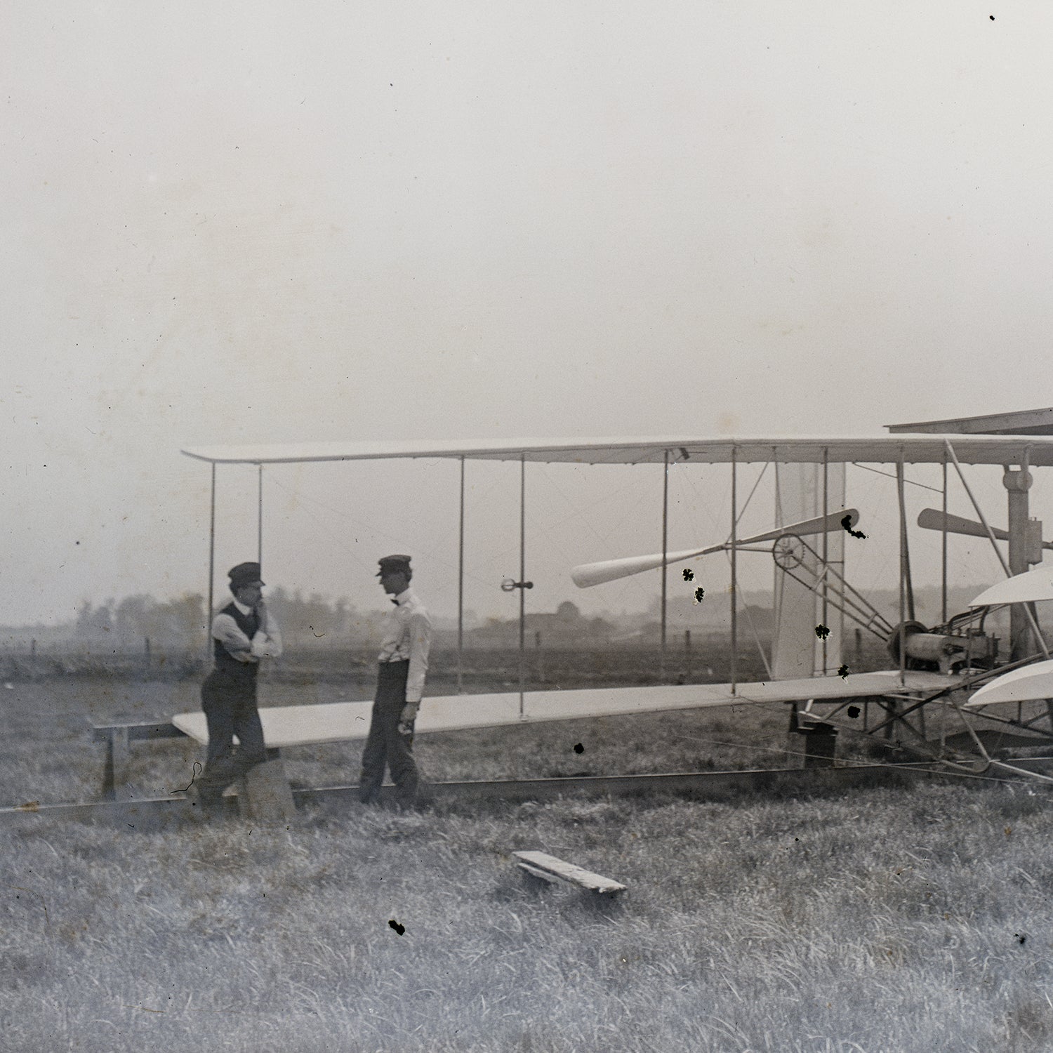Two men stand in front of a vintage biplane on a grassy field, evoking Early Aviation Art. The man on the left leans against the aircraft, while the man on the right stands upright, both gazing at the plane. The background is a foggy open space with a distant tree line, making it perfect for Historical Wall Decor. This scene is depicted in "The Wright Brothers Print.