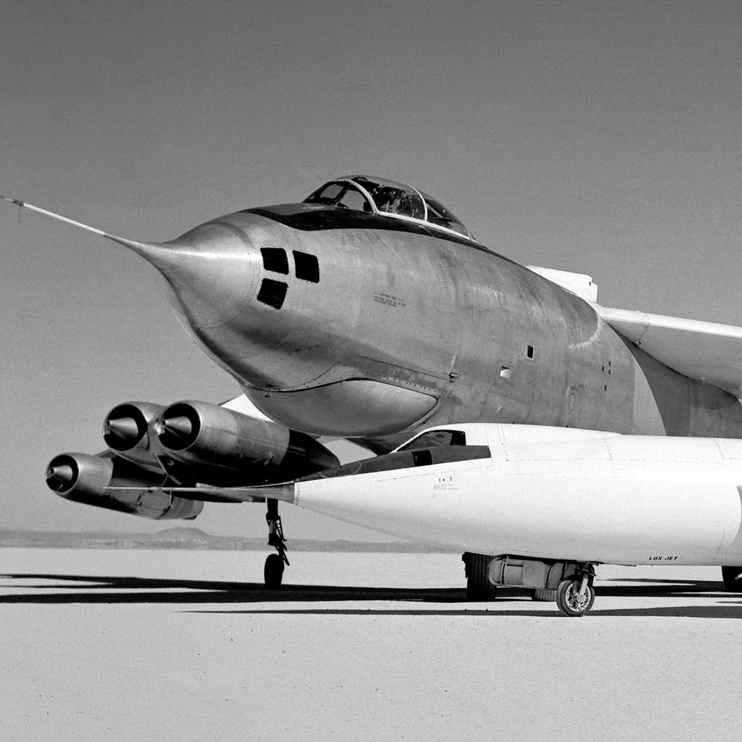 A close-up, black-and-white image from the "Douglas D-558-II and Boeing B-47A Stratojet Print | X Planes" collection captures the Boeing B-47A Stratojet parked on a flat, dry surface. The aircraft features a pointed nose, double cockpit, large armament pod on its underside, and two pairs of jet engines mounted on either side of its fuselage. The sky is clear.