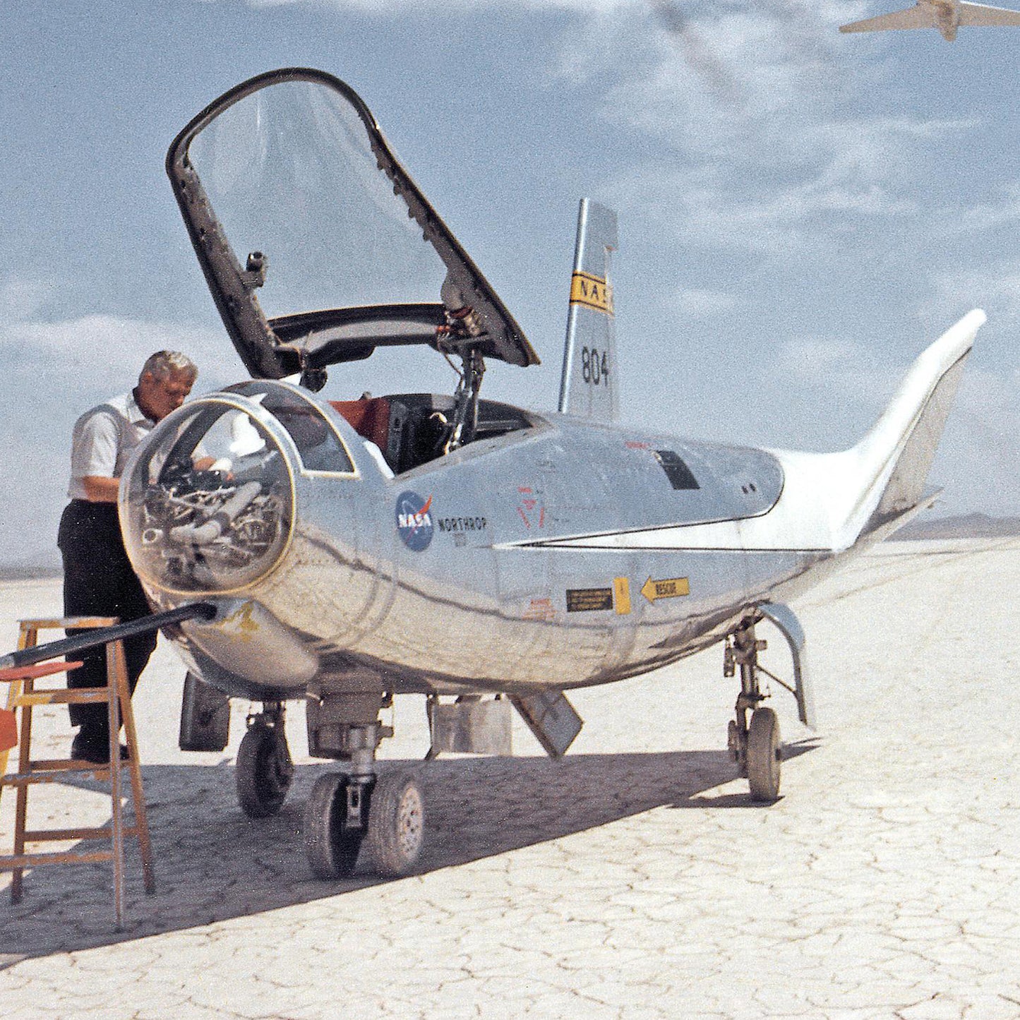 A man inspects NASA's silver-colored research aircraft, the HL-10 lifting body aircraft, designated with number 804, on a dry lakebed under a partly cloudy sky. The cockpit canopy is open, revealing the interior. Various technical markings and logos are visible on the aircraft's body.

Product: HL-10 on Lakebed with B-52 flyby Print