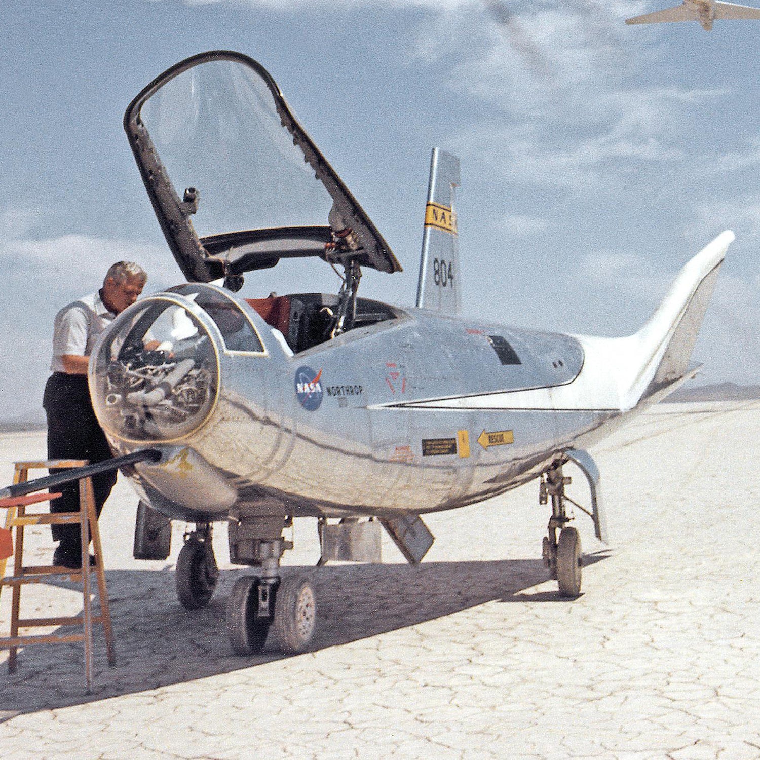 A man inspects NASA's silver-colored research aircraft, the HL-10 lifting body aircraft, designated with number 804, on a dry lakebed under a partly cloudy sky. The cockpit canopy is open, revealing the interior. Various technical markings and logos are visible on the aircraft's body.

Product: HL-10 on Lakebed with B-52 flyby Print