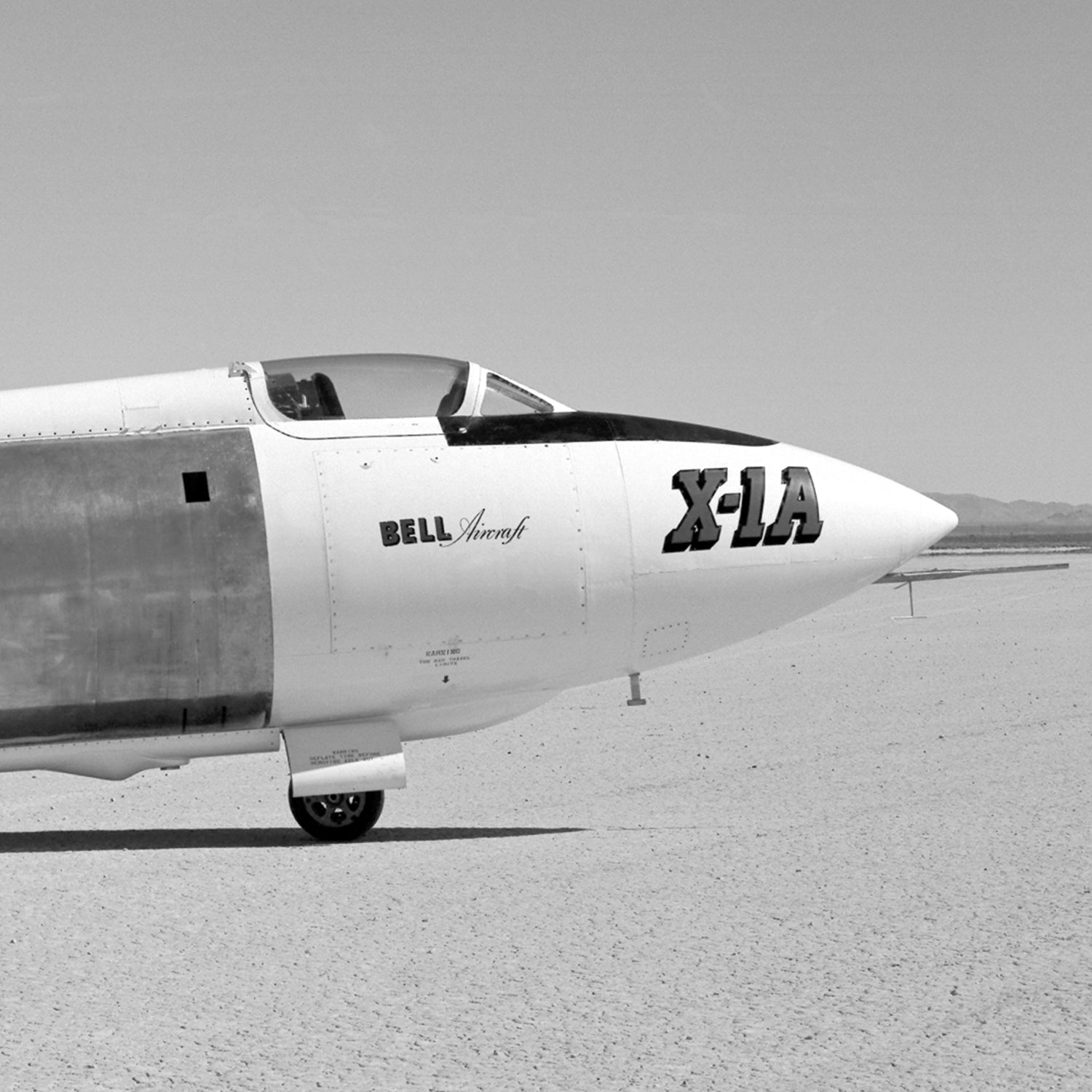 Black and white photograph of the X-1A Aircraft with Unpainted Fuselage Print from the X Planes series, highlighting its cockpit and front fuselage. The gleaming unpainted surface prominently displays "BELL Aircraft" and "X-1A" on its side. This groundbreaking vehicle for supersonic flight is captured resting on flat, barren ground with mountains in the distant background.