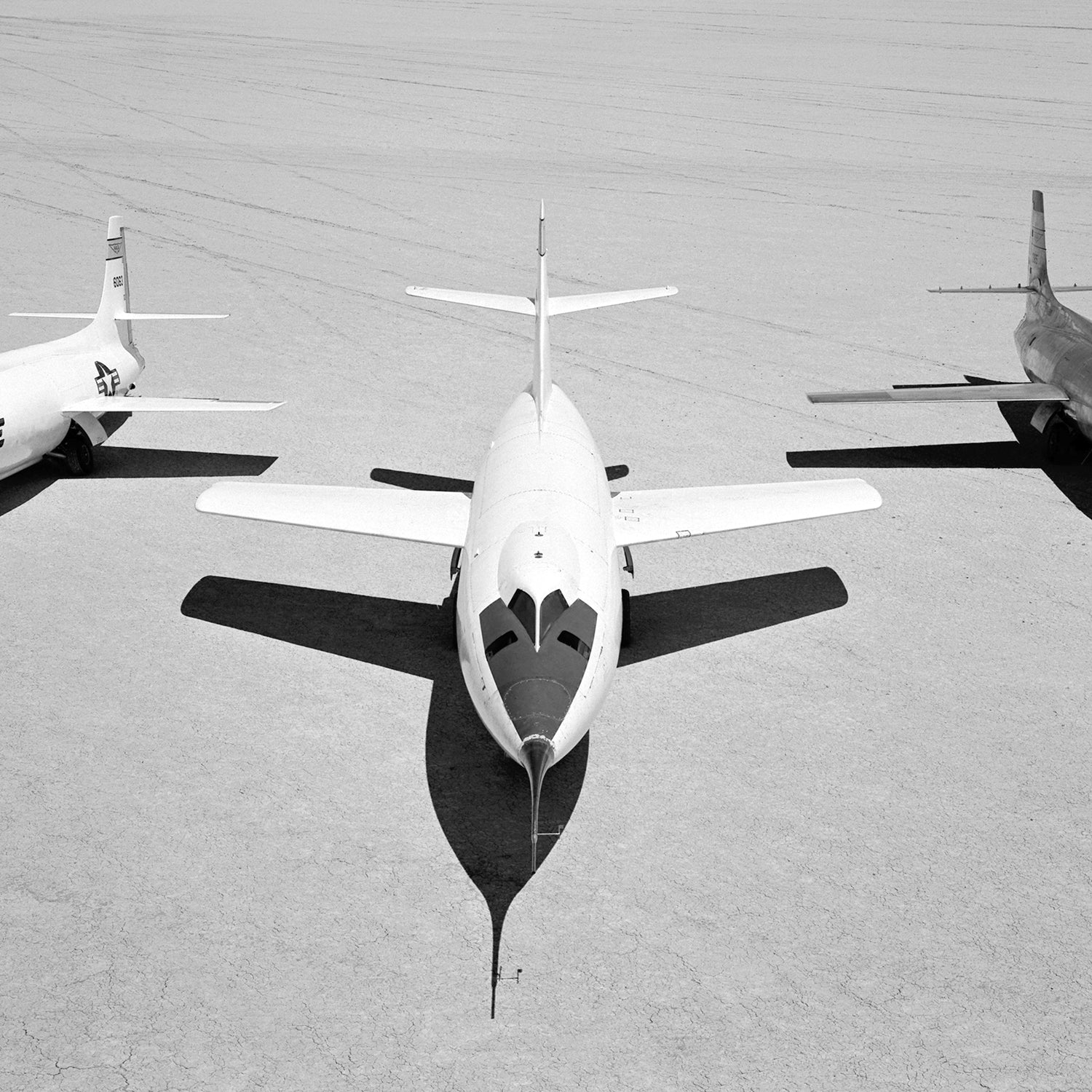 A black and white photo shows three vintage NACA aircraft parked on an expansive, dry lakebed. The central aircraft, resembling a sleek X-1B with its pointed nose, is flanked by two other planes—identified as the D-558-II and X-1E—with visible propellers and rudders. Their shadows are clearly cast on the ground.