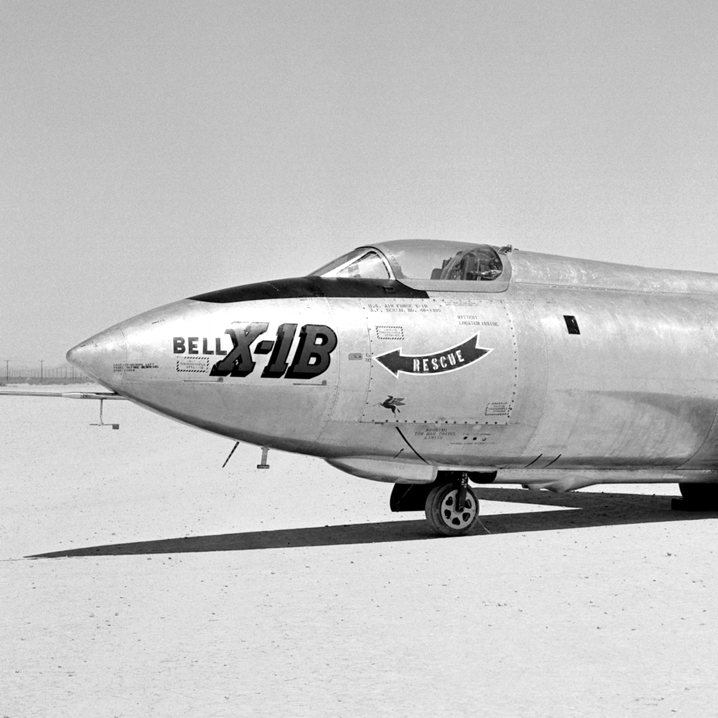 A black and white print titled "Bell Aircraft Corporation X-1B Rocket-Powered Aircraft | X Planes" captures the Bell X-1B, a pivotal rocket-powered aircraft in aviation history, on the ground. The plane's nose showcases large text reading "BELL X-1B" with a small "RESCUE" arrow pointing to a hatch. It is set in a desolate, flat area under a clear sky.