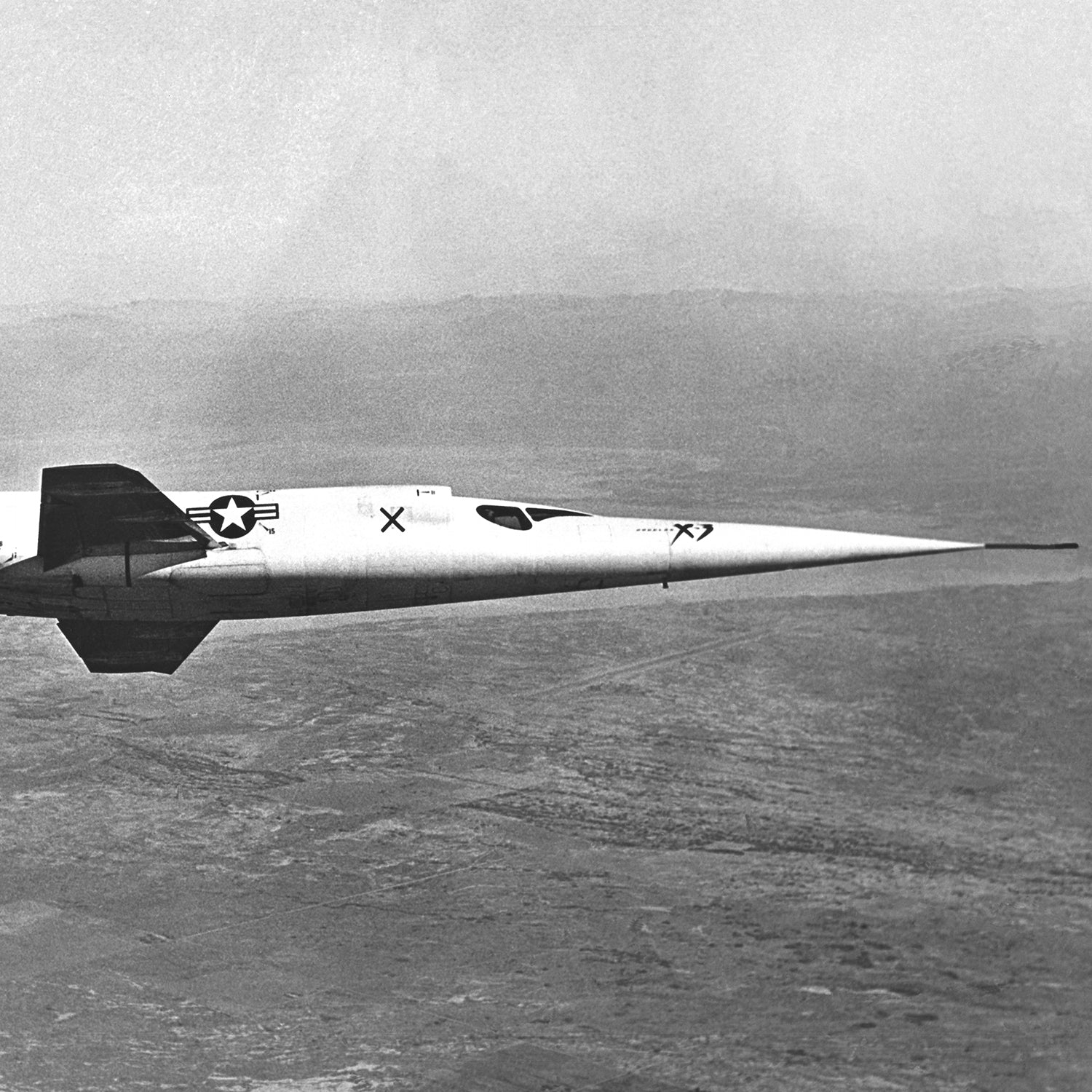 An in-flight photo depicts the sleek, high-speed experimental aircraft known as the Douglas X-3 Stiletto soaring through the sky. Its long, pointed nose and US Air Force insignia are clearly visible on the fuselage, set against a backdrop of a barren landscape and a hazy horizon.