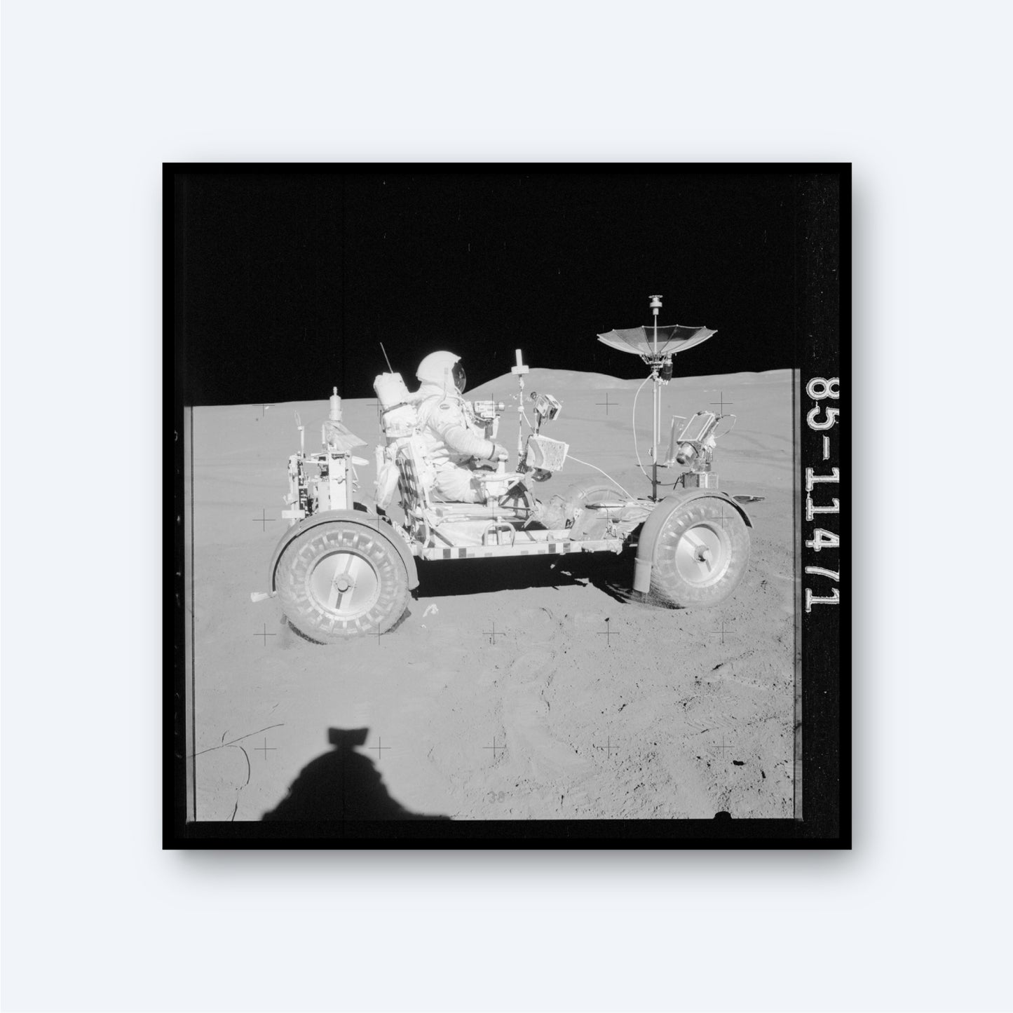 A black-and-white image of NASA Apollo 15 Astronaut David R. Scott driving the Lunar Roving Vehicle on the moon's surface. The vehicle, branded with NASA insignia, has large wheels and equipment attached. A dark, empty space is visible in the background, with the shadow of the photographer in the foreground.