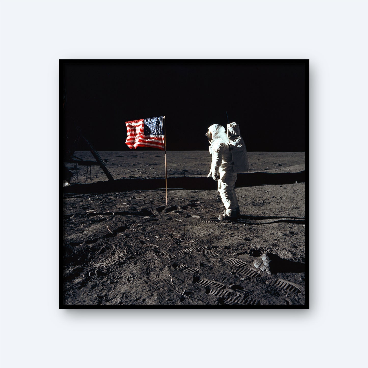 Buzz Aldrin, donned in an Apollo 11 space suit, stands on the Moon's surface beside The American Flag on the Moon with Buzz Aldrin Print. The ground is a dark, grainy carpet marked with footprints, and the pitch-black background emphasizes the stark and desolate lunar terrain.