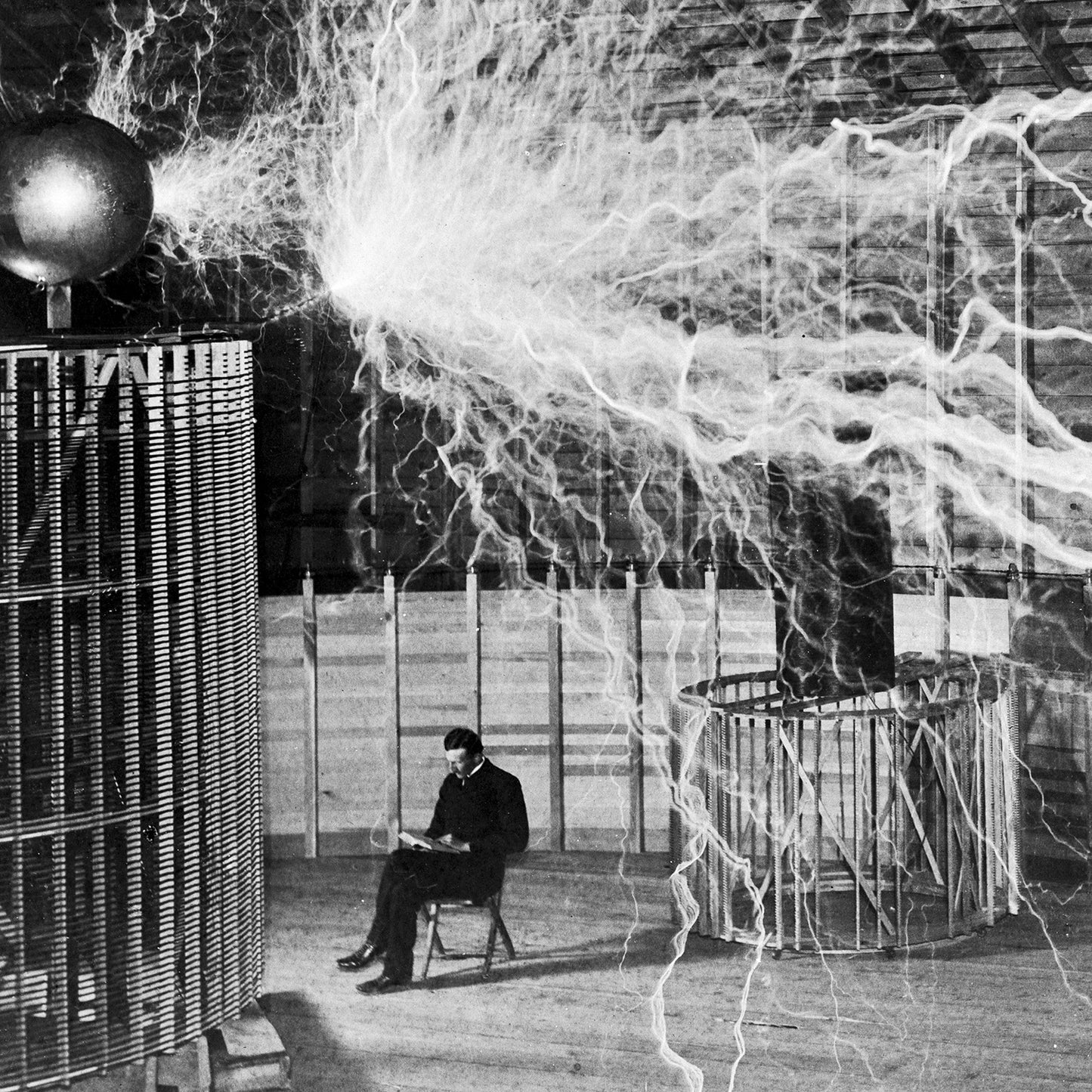 A black-and-white photo titled "Dr. Nikola Tesla in Laboratory Print" depicts a man seated on a chair in a large room, engrossed in reading papers. To his left, a massive Tesla coil emits numerous bright electric sparks and arcs across the room, representing the groundbreaking discoveries and technological advancements made by Tesla in his laboratory.