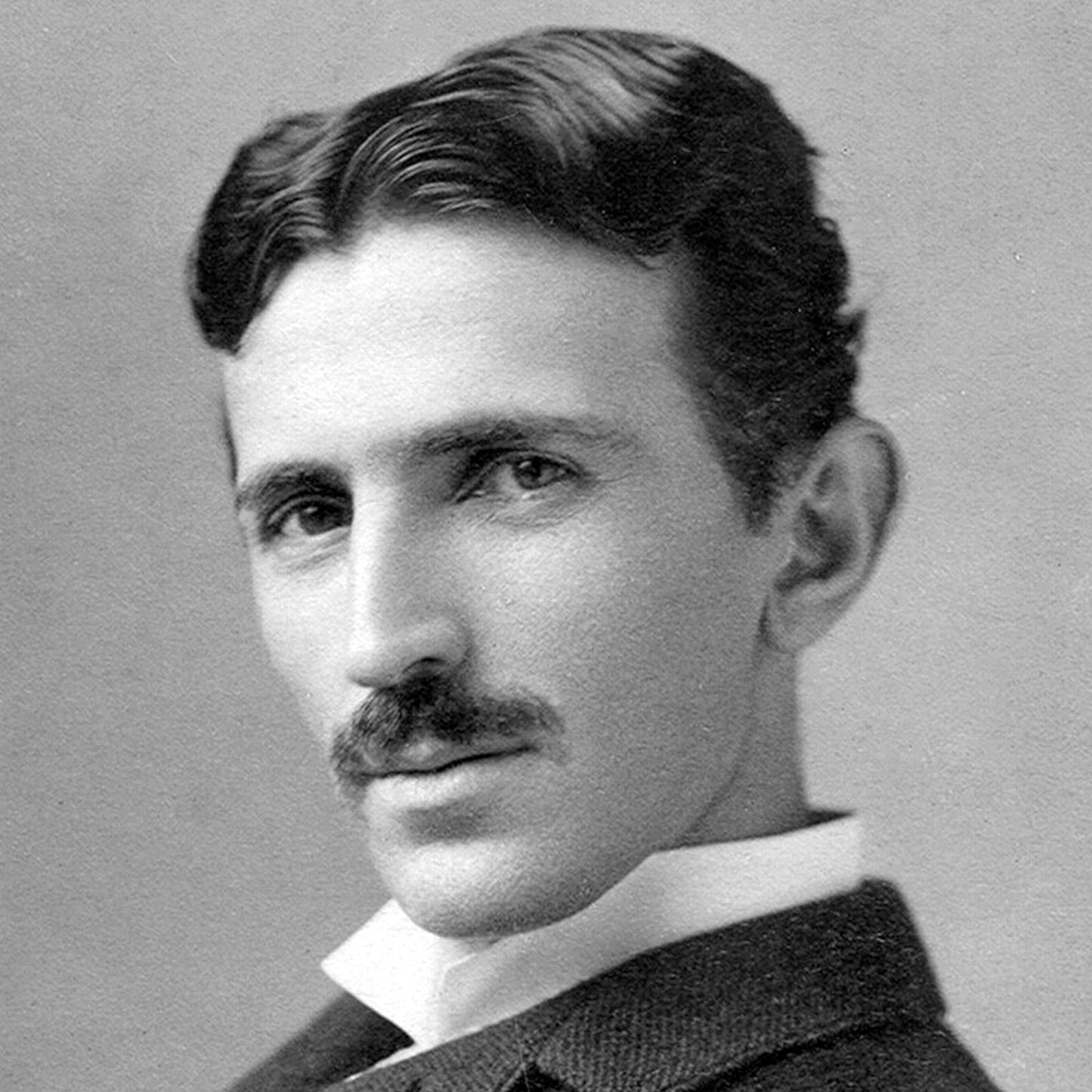 Black and white portrait capture of a man, styled with slicked-back hair and a short mustache. He is dressed in a suit complemented by a high-collared shirt. The subject's expression, serious yet thoughtful, reflects the iconic Nikola Tesla Print.