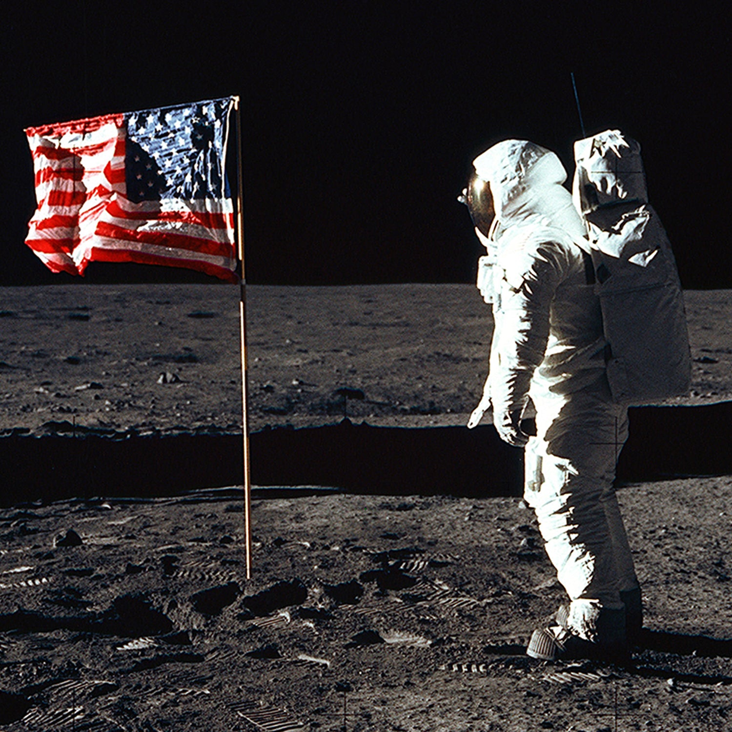 Buzz Aldrin, clad in a spacesuit, stands on the moon's surface, facing "The American Flag On The Moon With Buzz Aldrin" print. The moon's barren, dust-covered landscape is visible under a black sky—a silent testament to the historic Apollo 11 mission.