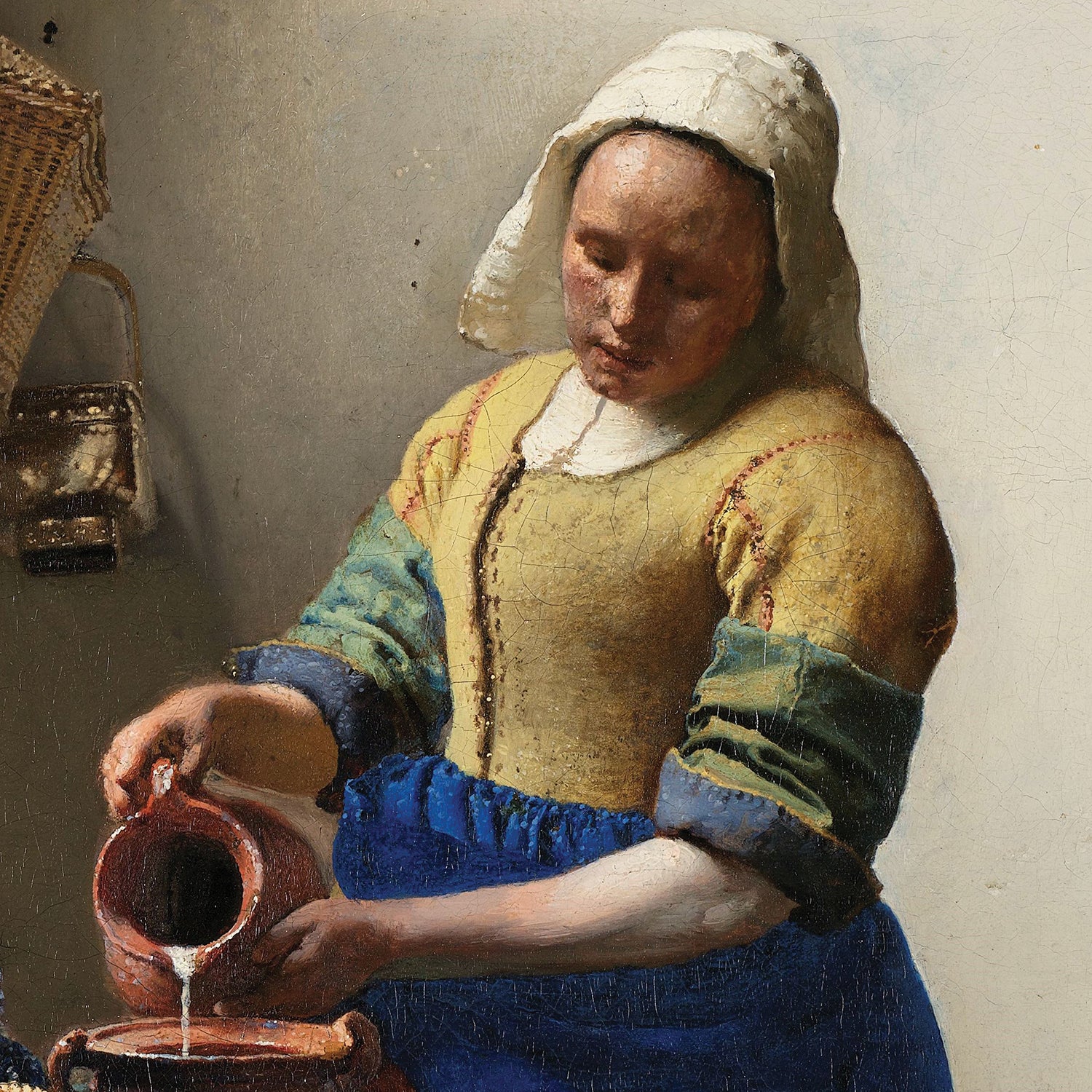 The Milkmaid by Johannes Vermeer depicts a woman in a white headscarf, yellow bodice, blue apron, and green-and-blue sleeves, pouring milk from a red jug into a bowl held by her left hand. In the background, a basket and loaf of bread are visible, highlighting the Dutch Golden Age style characteristic of Vermeer's work.