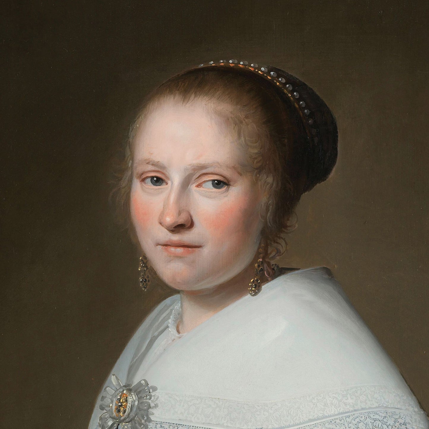 A painting titled "Portrait of Maria van Strijp by Johannes Cornelisz," featuring a woman with light skin and red cheeks. She is dressed in a white shawl adorned with a brooch at her chest, has her hair pulled back, and wears a decorative headband with pearls and dangling earrings. The woman gazes slightly off to the left against a dark background, evocative of the Dutch Golden Age.