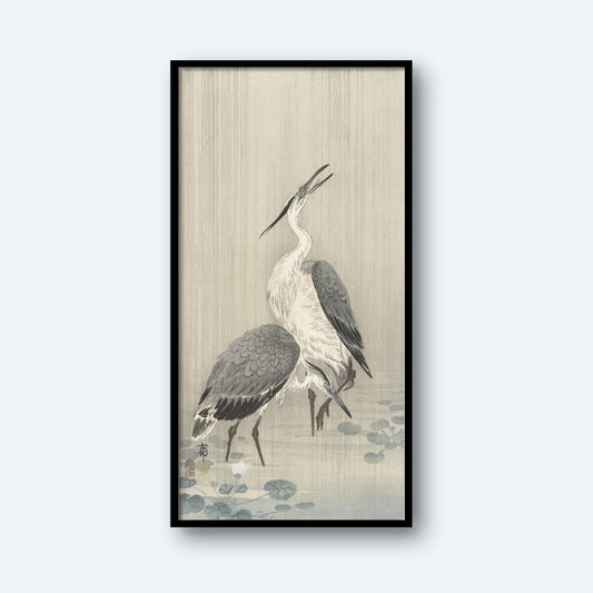The vertical framed artwork titled "Two Herons in the Rain by Ohara Koson | Vintage Japanese Print" captures two herons standing among lotus leaves. One heron has its beak pointed upwards, while the other rests with its beak pointed down. The background features a soft gradient of light beige and gray tones, creating a serene atmosphere reminiscent of an authentic Ohara Koson print.