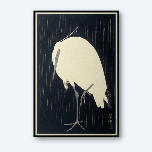 The vintage Japanese print "Egret in the Rain" by Ohara Koson captures a minimalist artwork reminiscent of traditional woodblock printing. It features a white egret standing on one leg against a dark background, with finely detailed feathers that contrast beautifully with the simple backdrop. The piece is adorned with Asian script in the lower right corner and is framed in black.