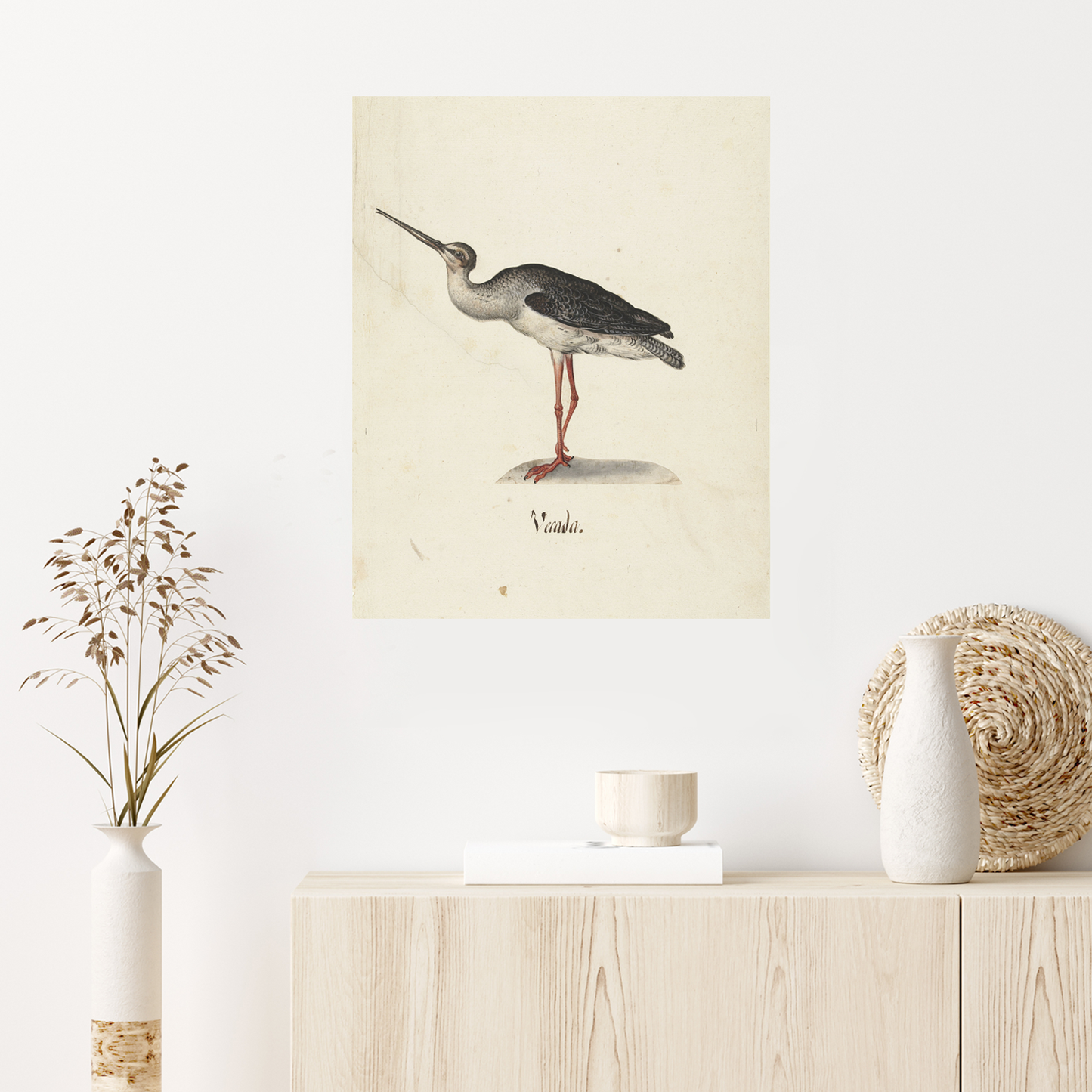A minimalist room with a wooden sideboard features a framed drawing of a heron on the wall. The sideboard is decorated with a white vase holding dried flowers, a small bowl, and a woven basket lid leaning against the wall. Adding to the charm is some Black Rider Vintage Print wall art.